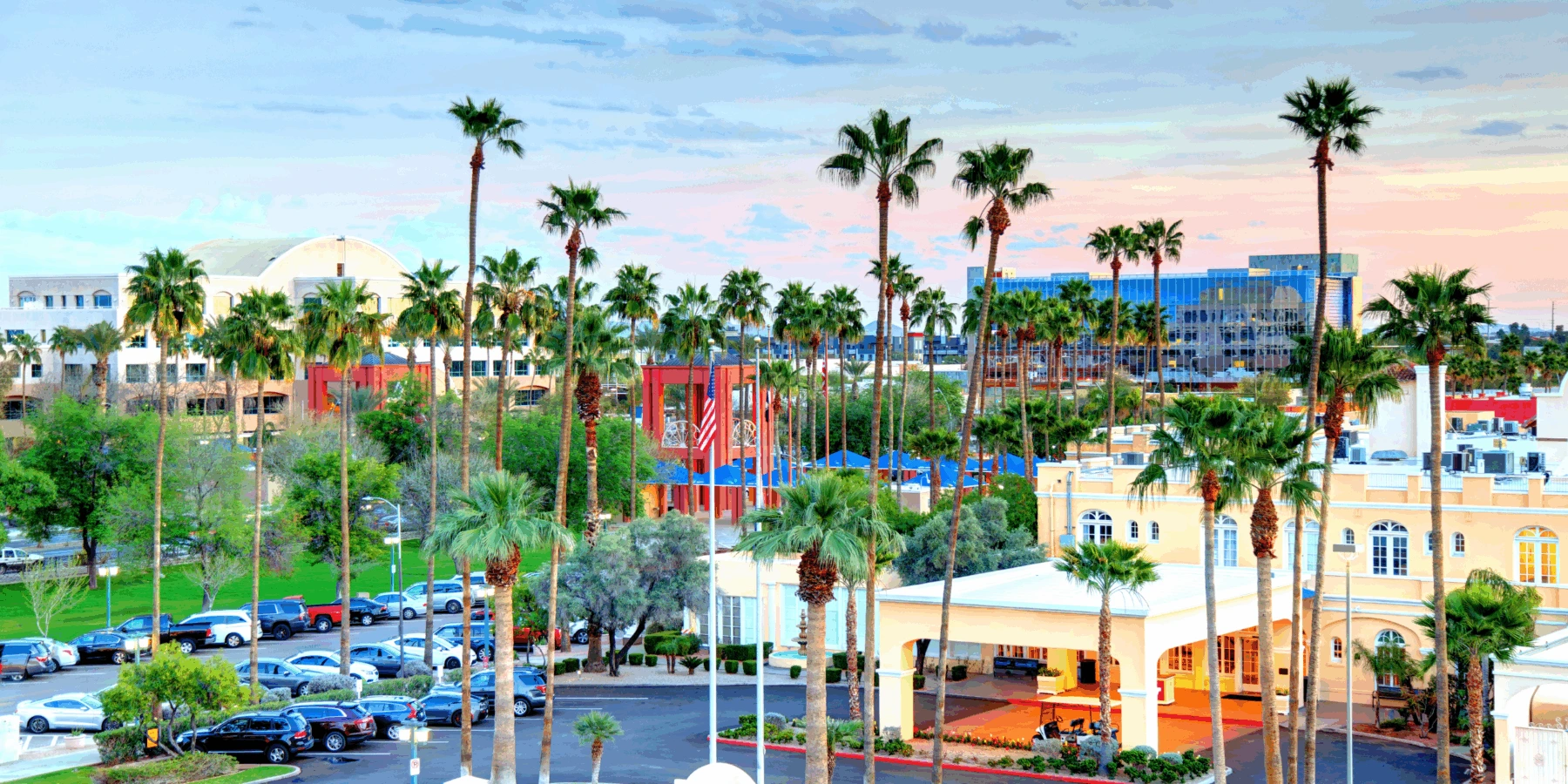 Sunny resort landscape with tall palm trees, a parking lot, and corporate buildings under a sunset sky.