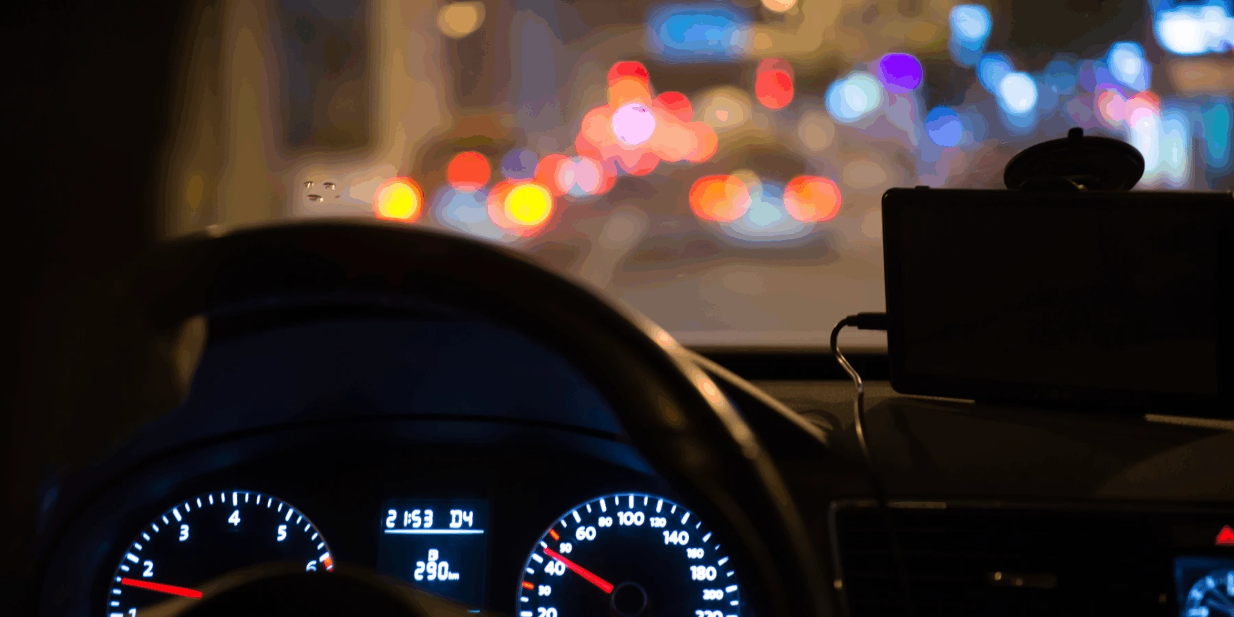A steering wheel of a car driving at night, showing an illuminated dashboard and a phone mounted on the dash against a background of blurred city traffic lights.