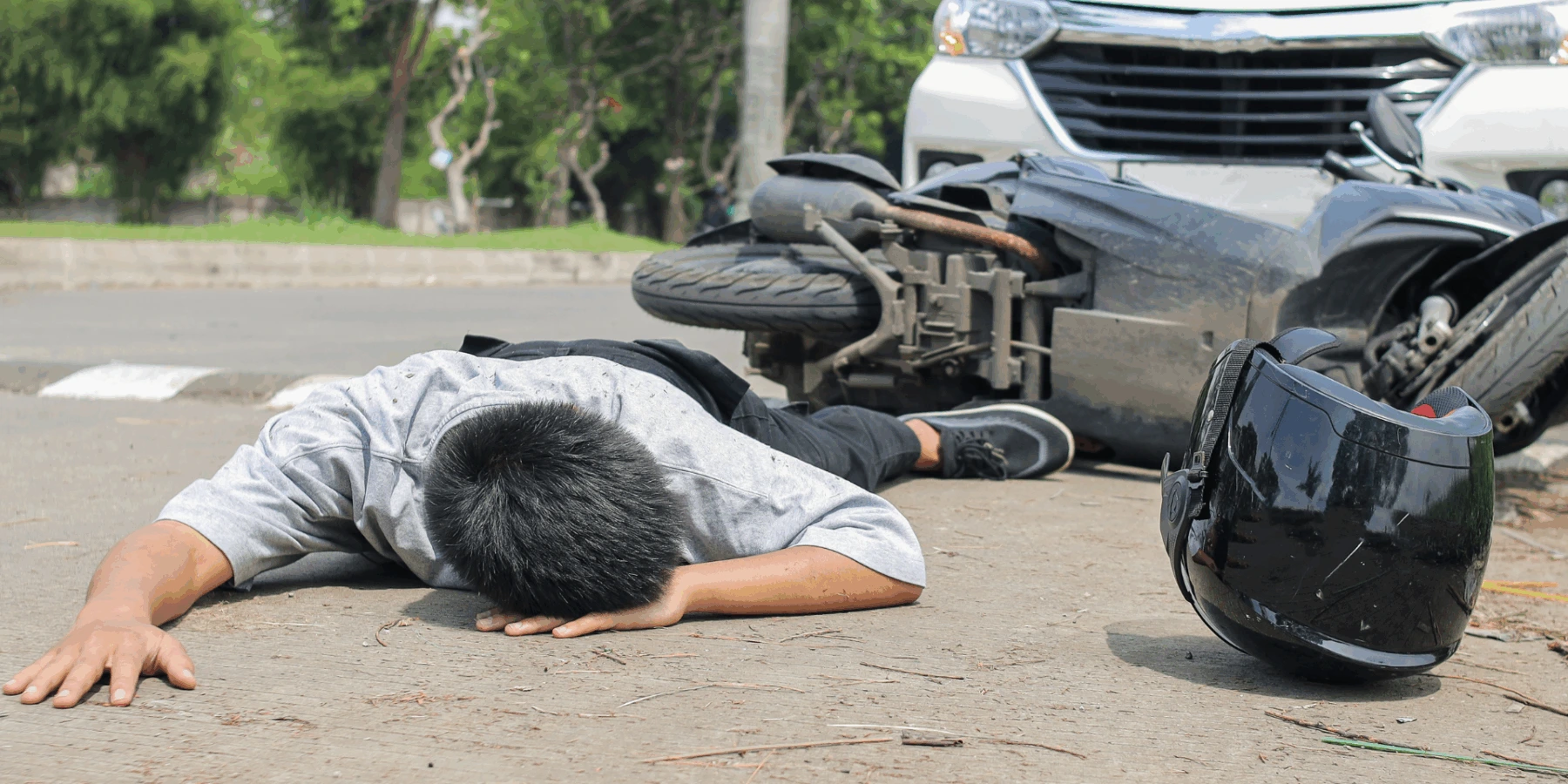 A motorcycle accident scene with a person lying on the road near a fallen bike and a white car.