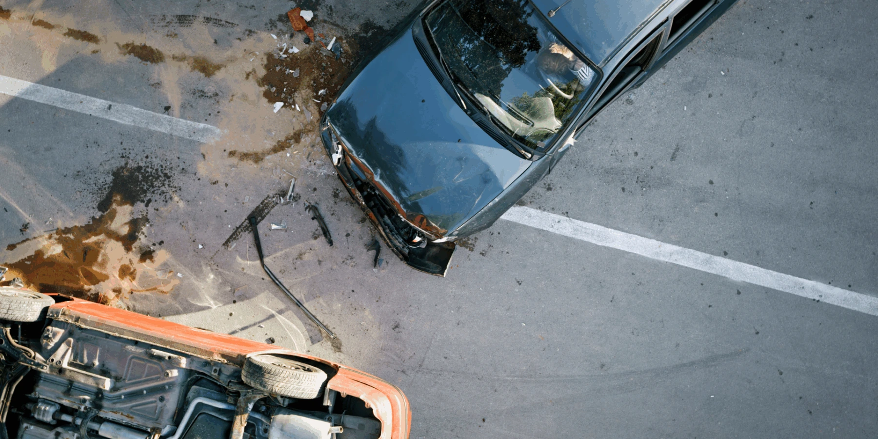 A car accident scene showing a dark sedan with front-end damage and a red vehicle overturned on its side, surrounded by debris and fluid spills on the asphalt.