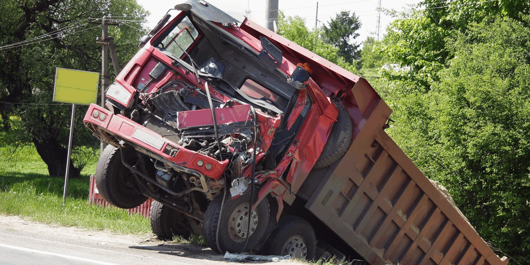 A red dump truck with a heavily damaged front cab tilted upward and backward after a collision on a roadside.