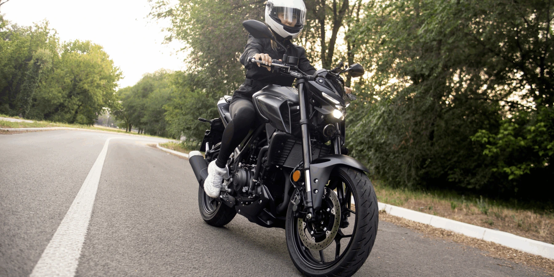 A motorcyclist wearing a white helmet and black gear riding a black motorcycle on a curved paved road lined with trees.
