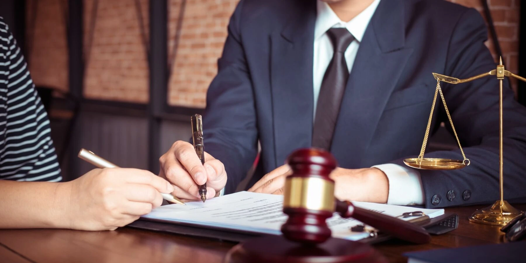 An attorney in a suit sitting at a desk with a client, featuring a legal gavel, and scales of justice.