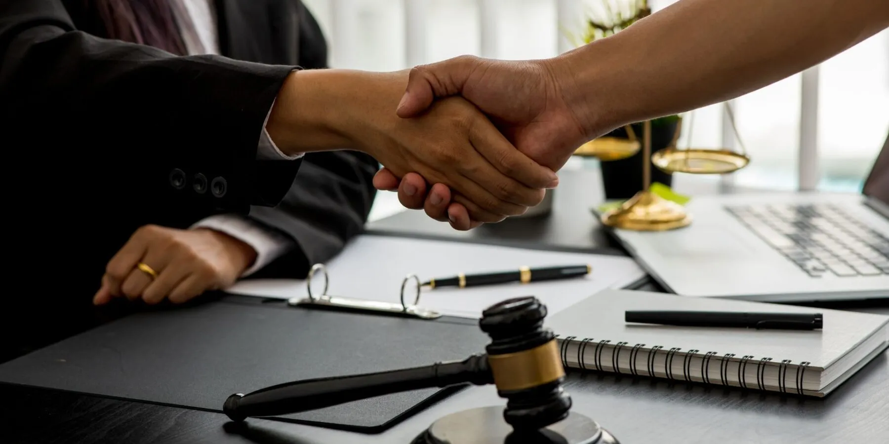 Two professionals handshake over a legal desk with a gavel and scales of justice.
