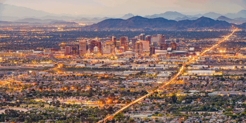 A sprawling city skyline at dusk, with glowing city lights, lit highways, and mountains in the background.