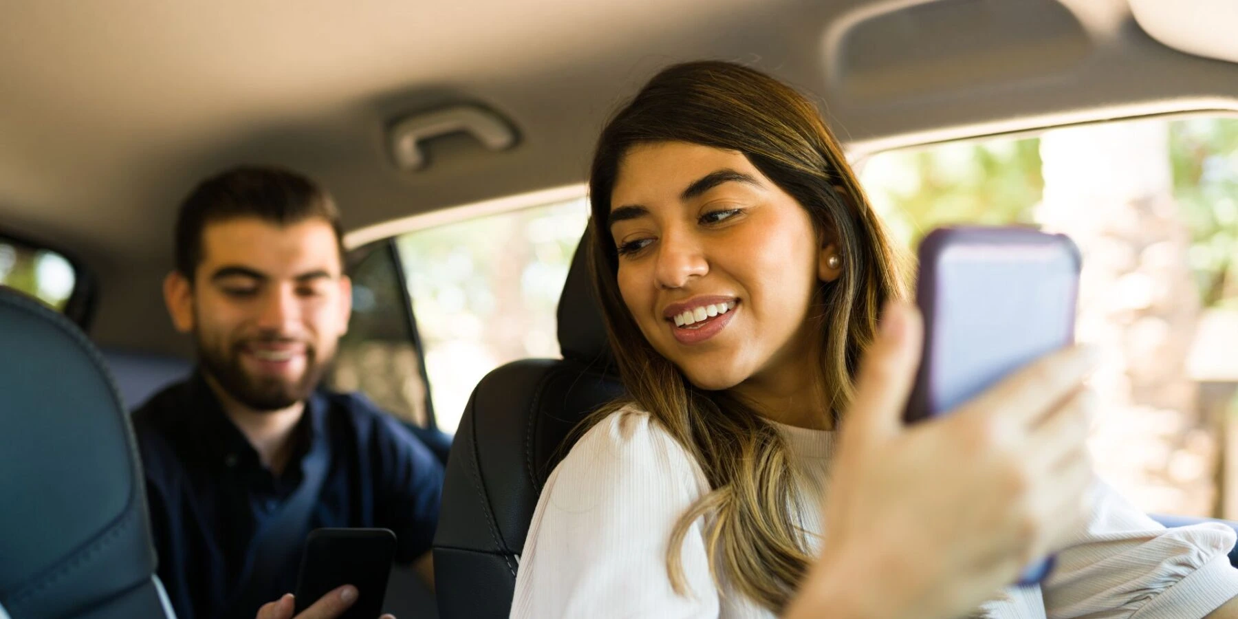 A smiling woman in the passenger seat of a car holds up her smartphone, looking toward the device.
