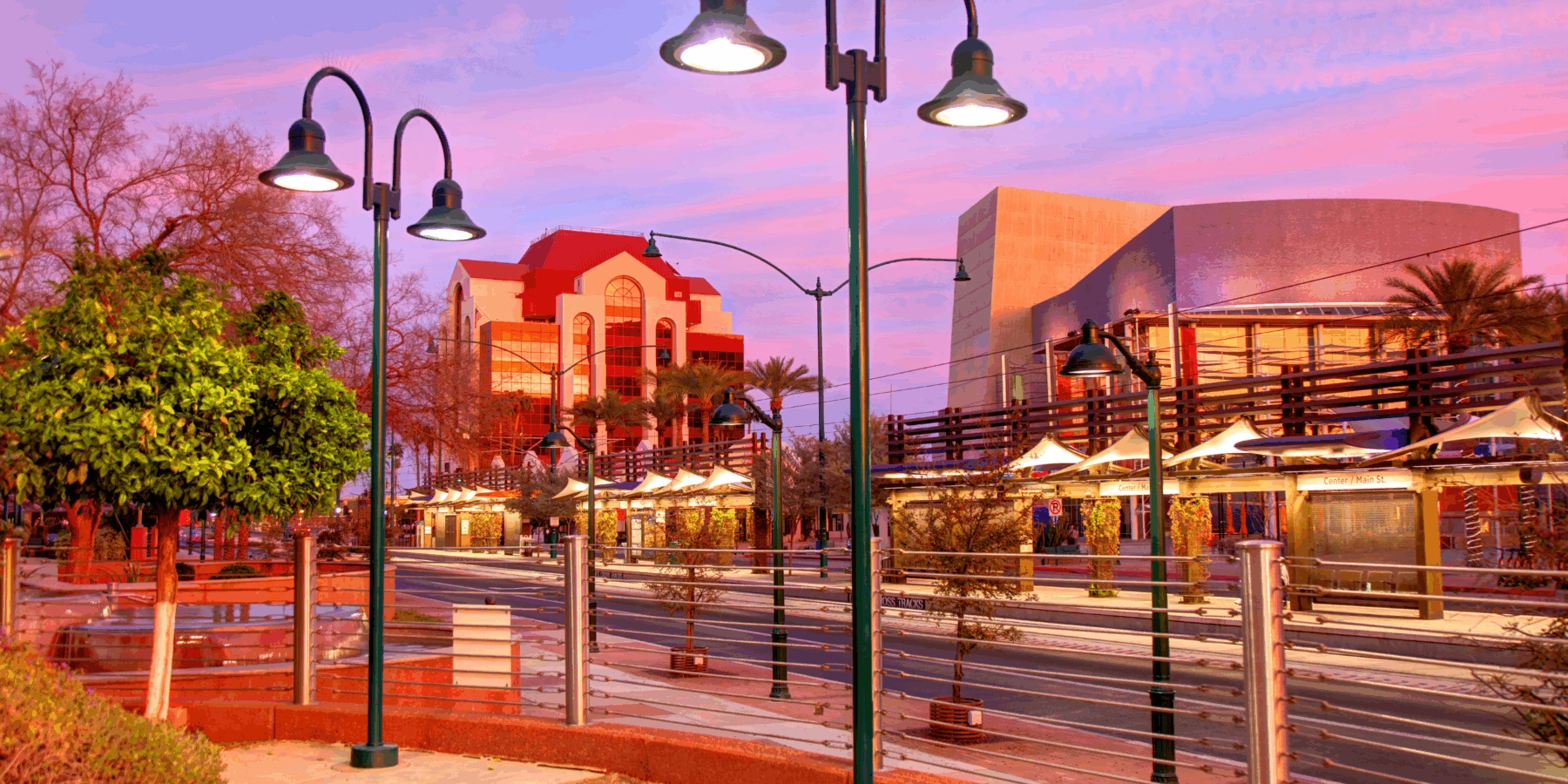 A city transit station and modern buildings under a vibrant purple and pink sunset sky, framed by lit street lamps.