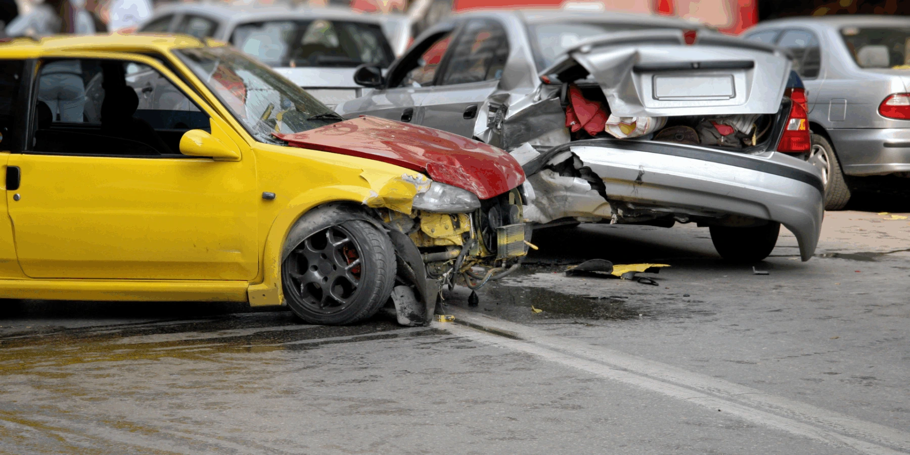 A yellow car with front-end damage and a silver car with a crushed rear bumper following a multi-vehicle collision on a city street.