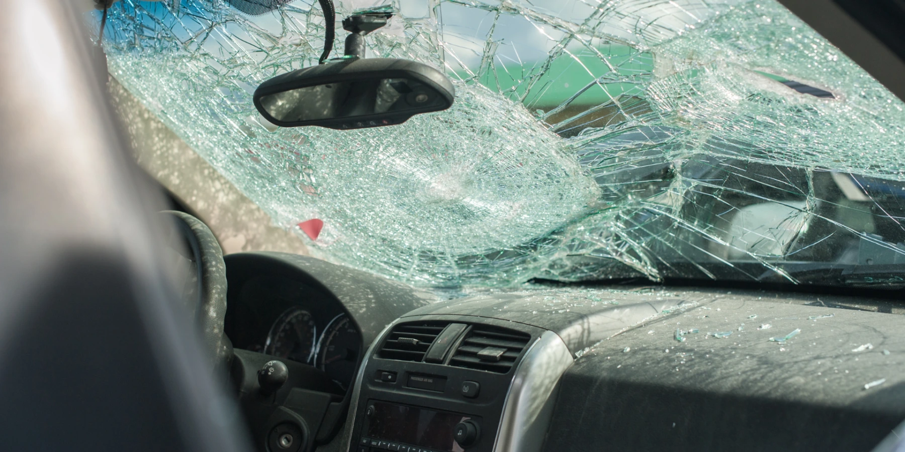 A car showing a severely shattered windshield with a prominent impact point, with glass shards scattered across the dashboard.