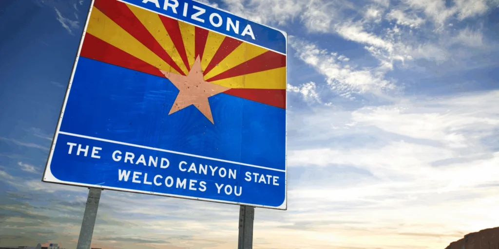 Arizona welcome sign with the state flag and "The Grand Canyon State Welcomes You" against a bright sky.Arizona welcome sign with the state flag and "The Grand Canyon State Welcomes You" against a bright sky.
