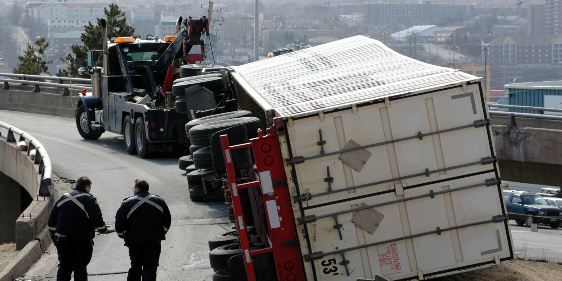 A large white shipping container trailer is overturned on its side across a highway ramp.