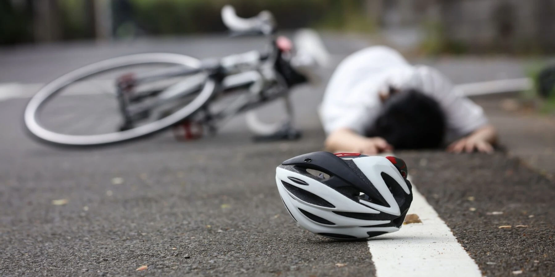 A person lies on an asphalt road next to a fallen bicycle and a discarded helmet.