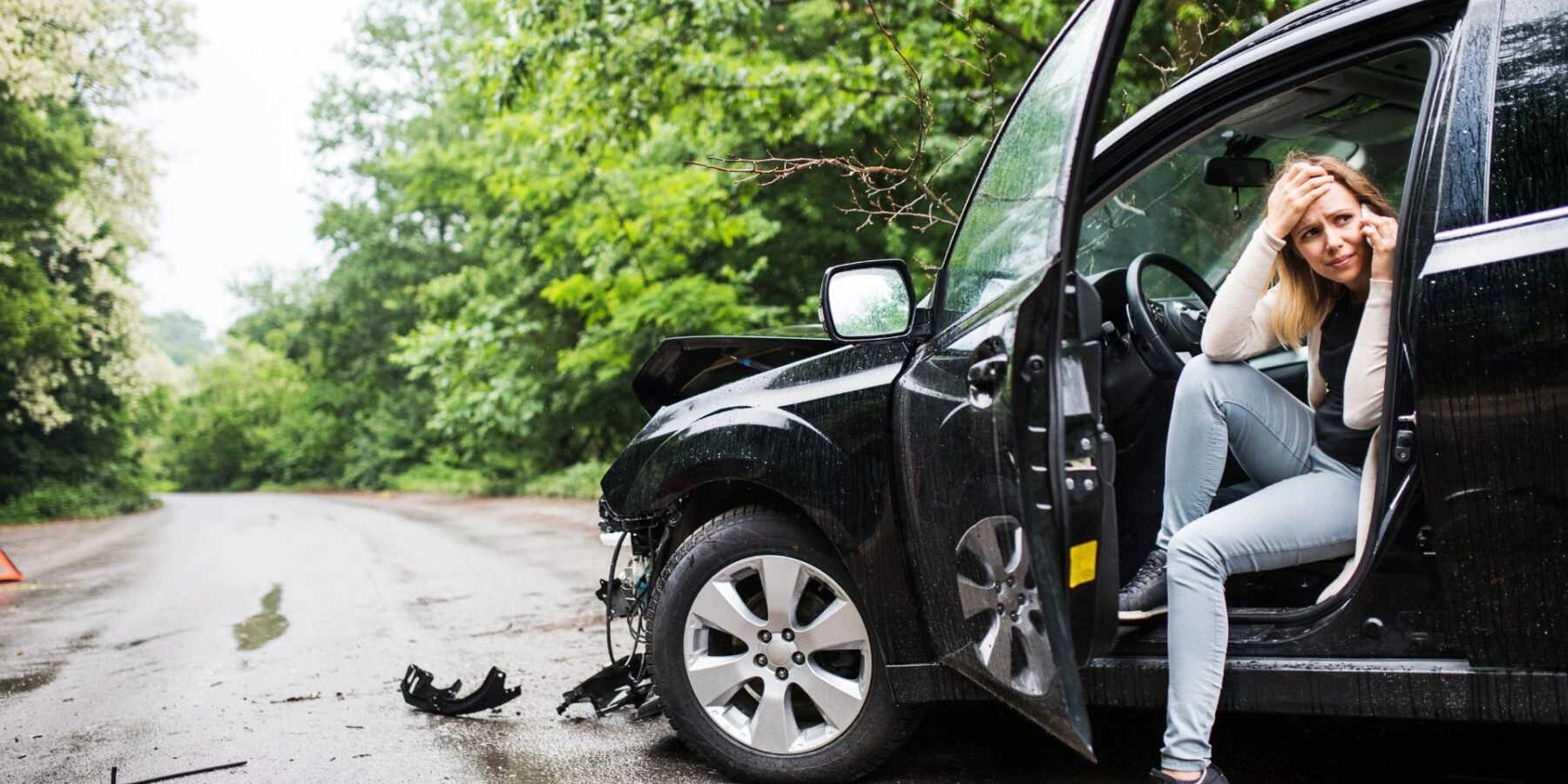 A distressed woman talks on a phone while sitting in the doorway of a damaged black car.