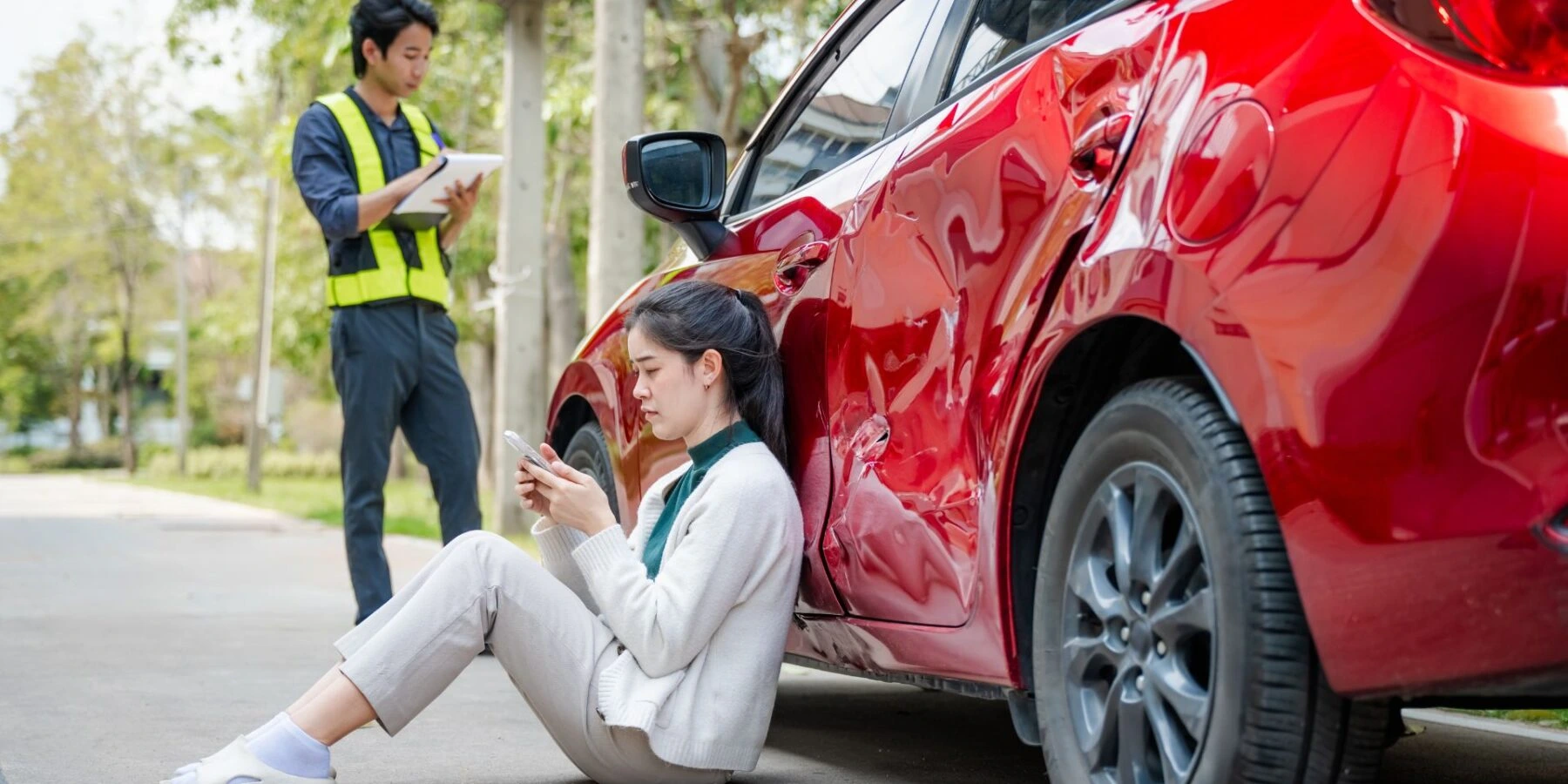 Woman on phone by a dented red car while a man in a safety vest takes notes.