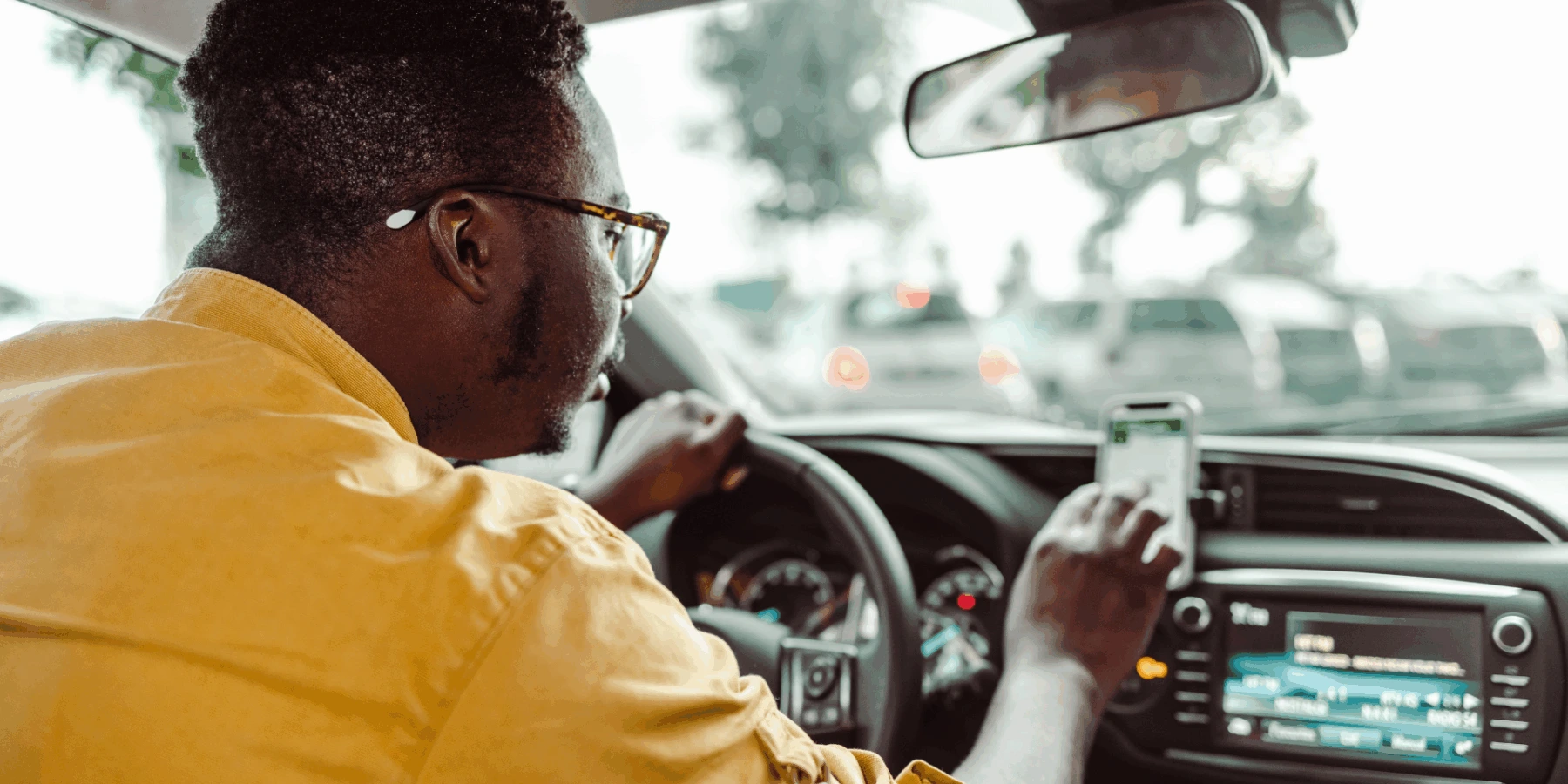A man in a yellow shirt and glasses touches a dashboard-mounted smartphone while driving.
