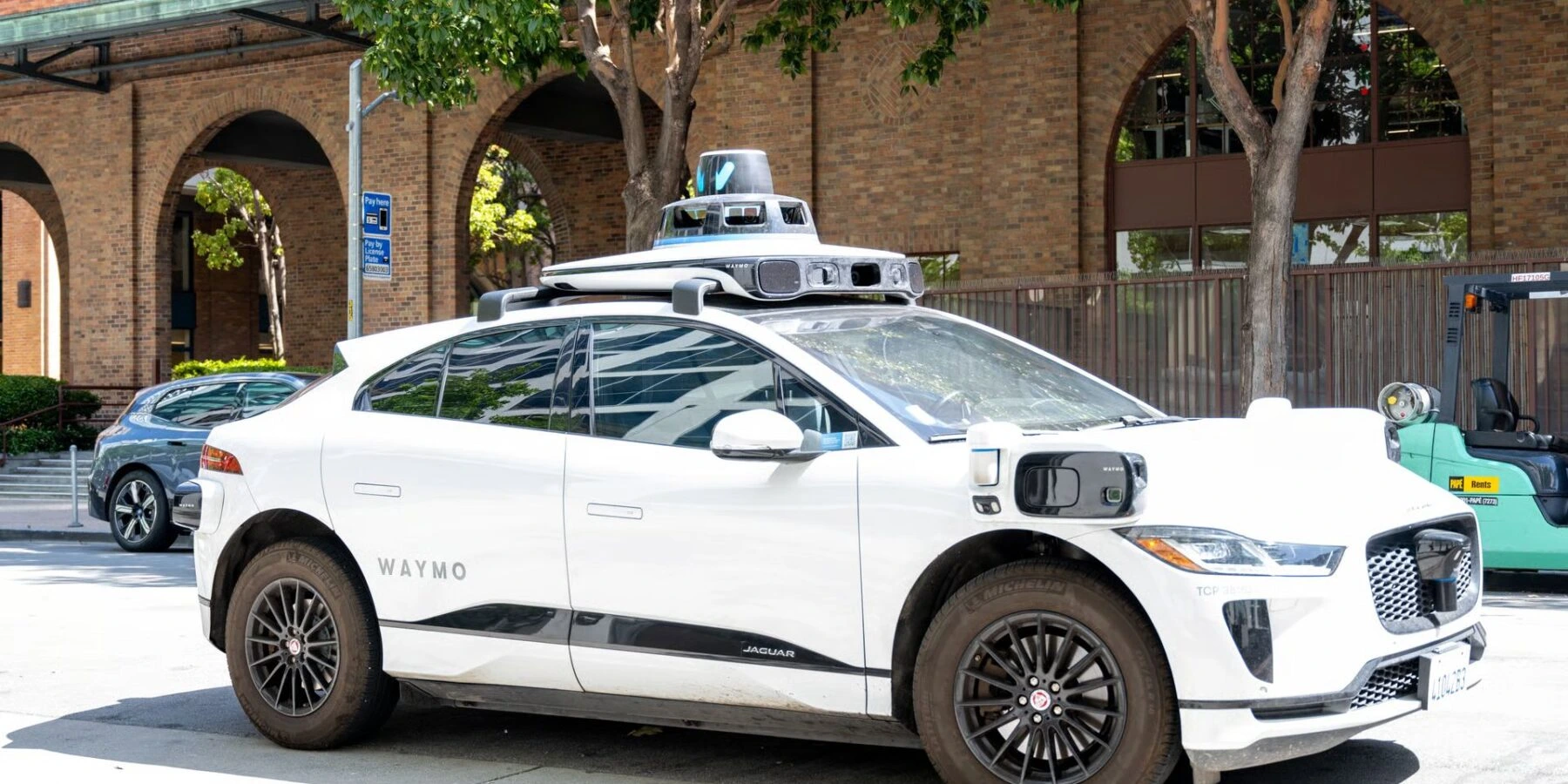 A white Waymo self-driving Jaguar I-PACE is parked on a city street, equipped with a prominent LiDAR sensor array and cameras on its roof and sides.