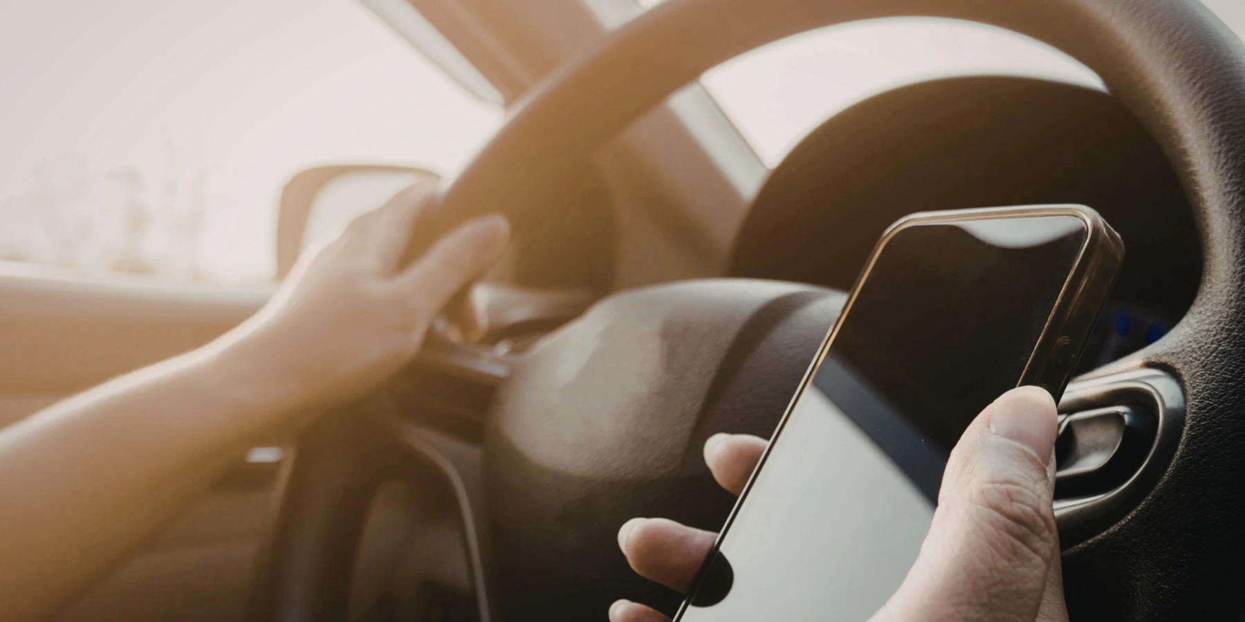 A driver's hand holding a smartphone while the other hand is on the steering wheel, illustrating distracted driving inside a car.