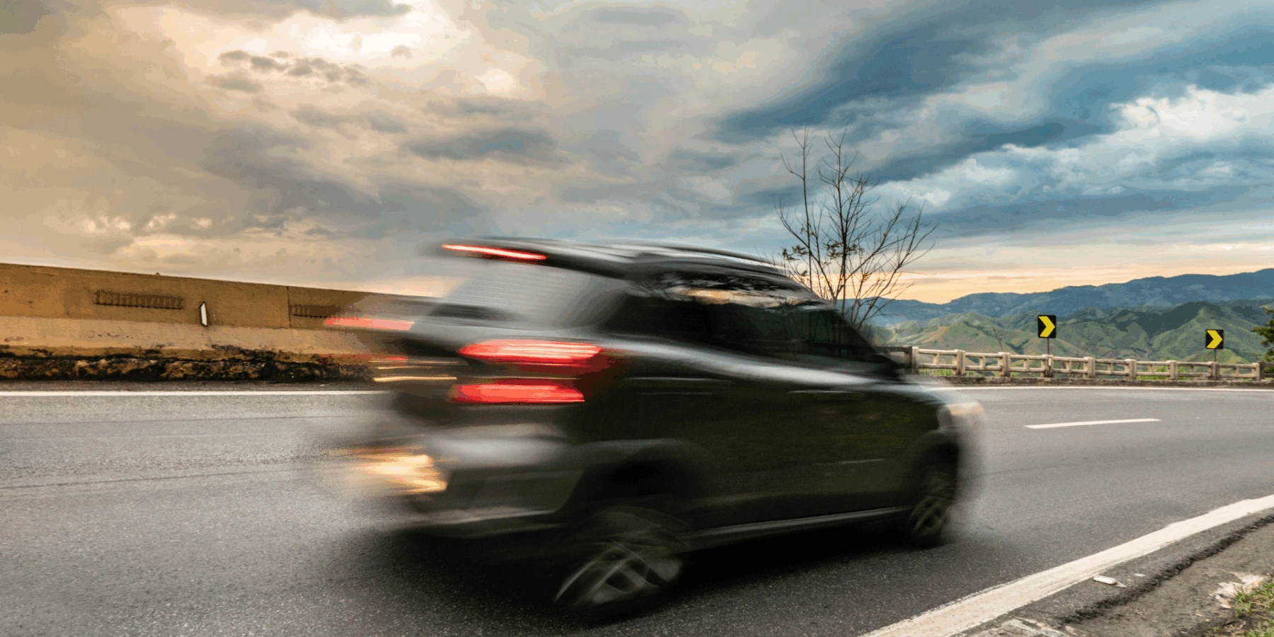 A motion-blurred dark SUV driving along a winding mountain highway under a dramatic, cloudy sky with green hills.