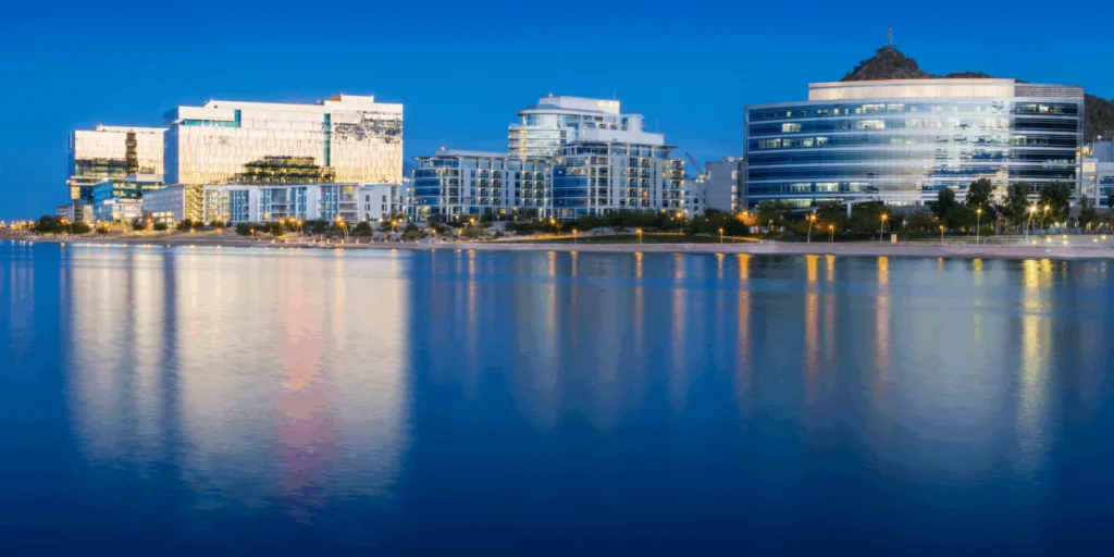 A row of modern, lit office and residential buildings along a waterfront at twilight, with their lights reflecting on the calm blue water.