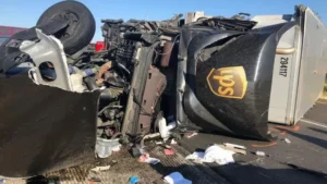 An overturned brown UPS semi-truck lies on its side with severe cab and engine damage.