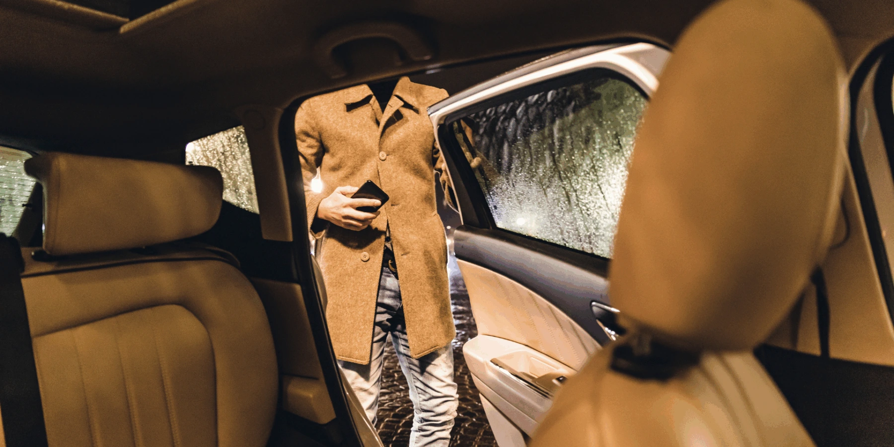A man in a tan overcoat holding a smartphone as he prepares to enter the back seat of a car at night during a rainstorm.