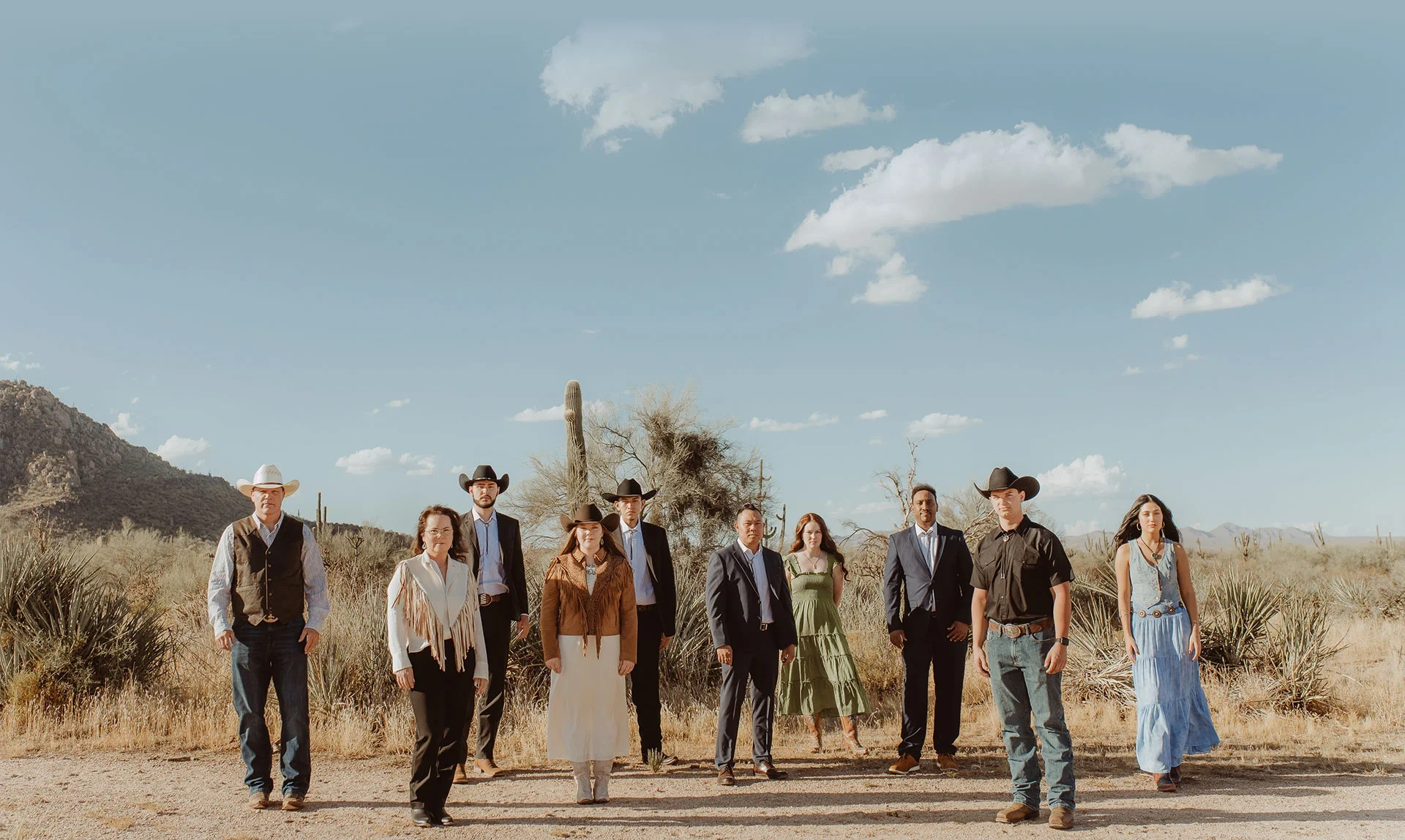 A group of ten people in western attire and cowboy hats poses in a desert landscape.